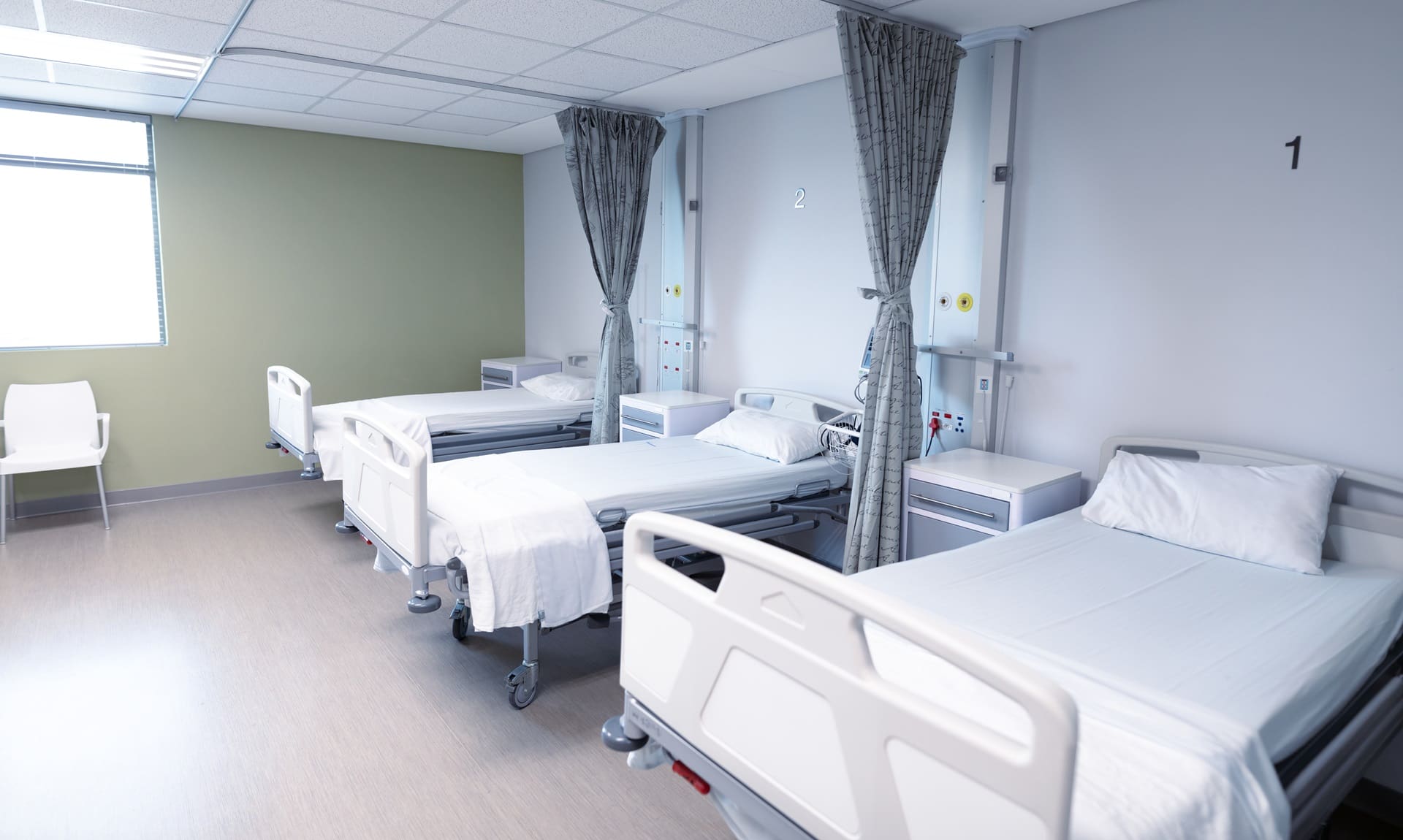 Hospital room with three empty beds, white sheets, and privacy curtains. Natural light enters through a window, illuminating the clean, organized space.