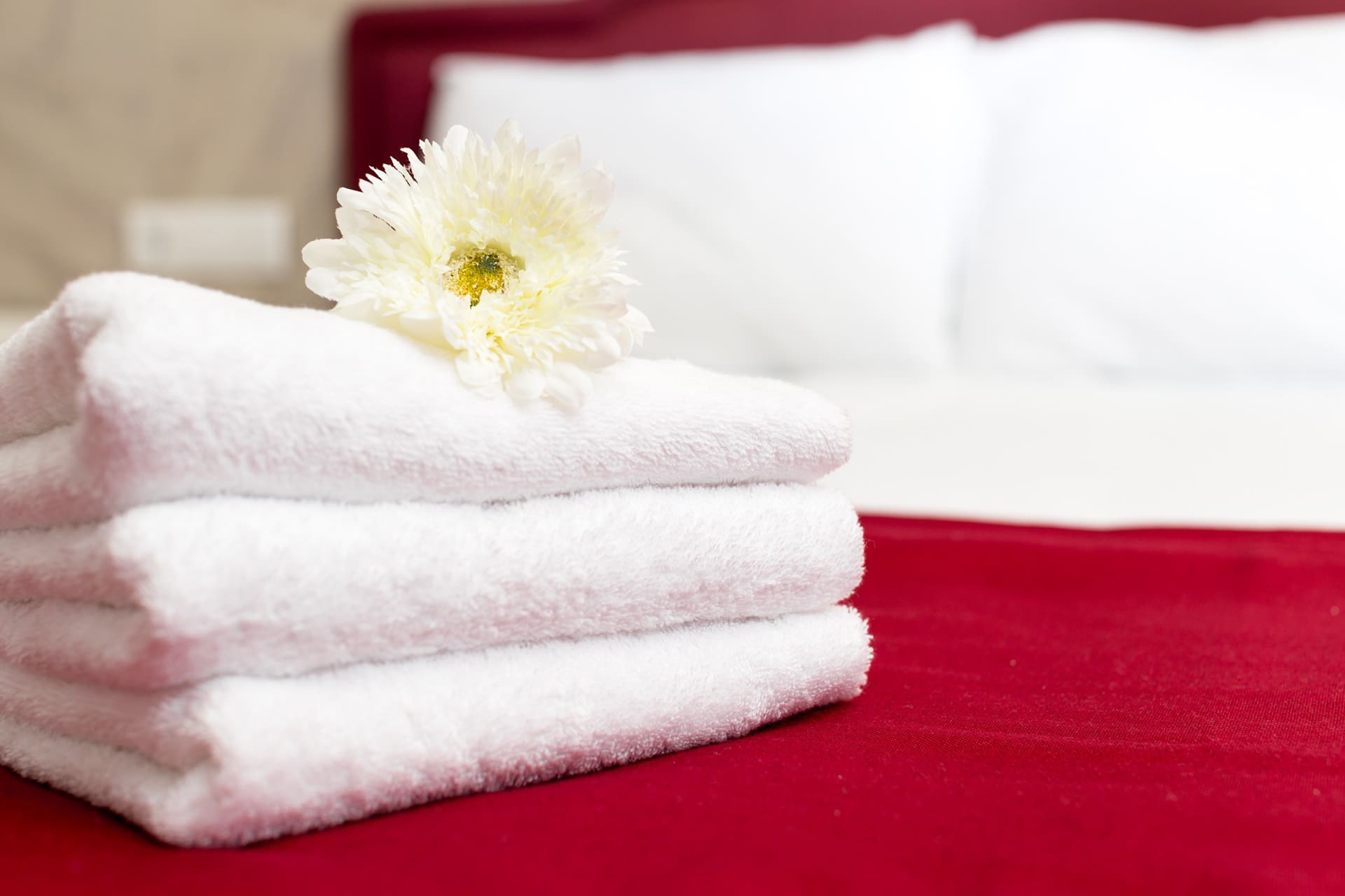 A stack of three folded white towels with a white flower on top, placed on a red bedspread with white pillows in the background.