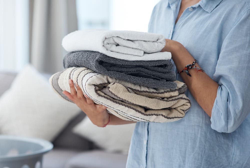 A person holding a stack of freshly folded towels, featuring white, gray, and striped patterns, showcases the meticulous care typical of a quality laundry service in a cozy living room.