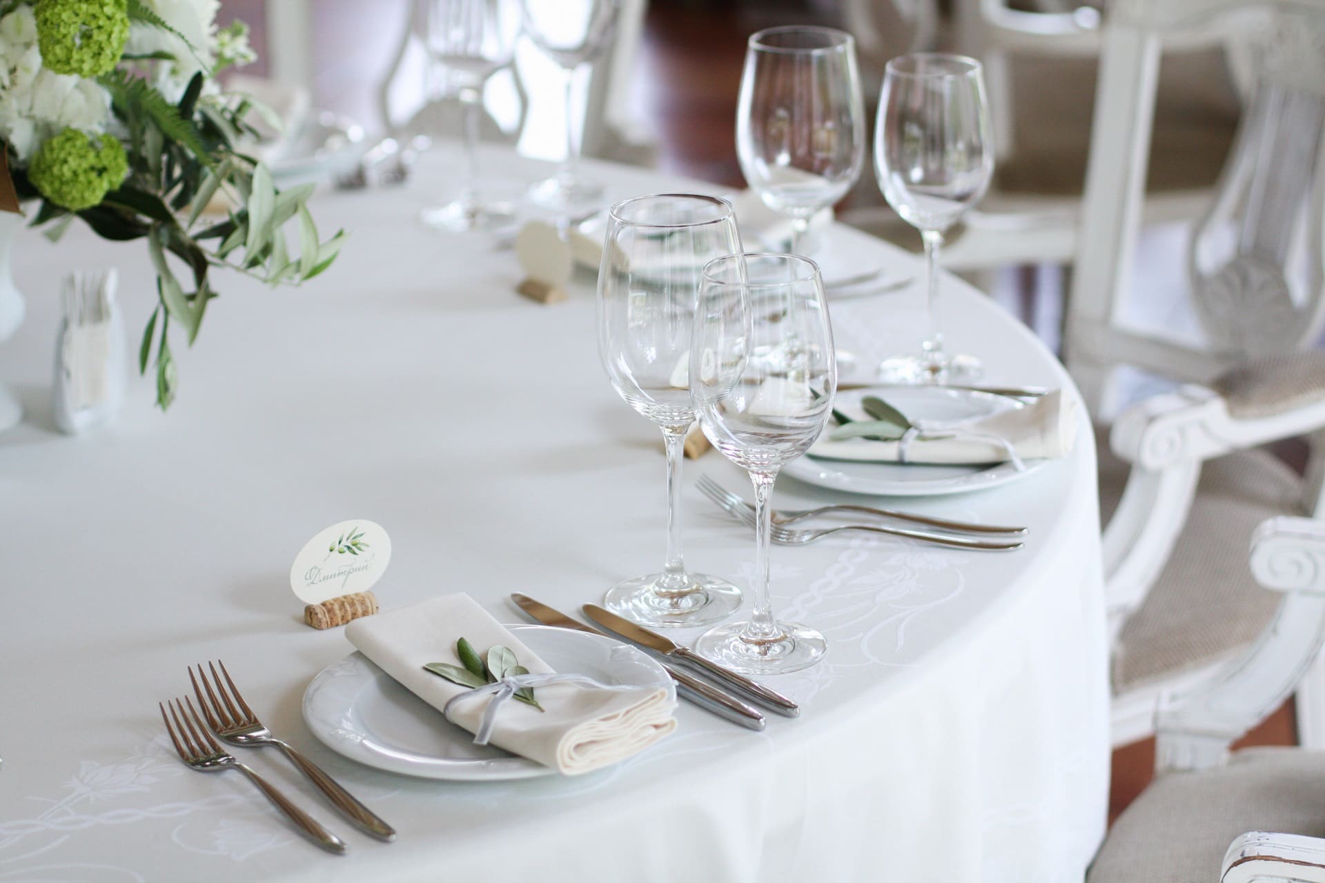 Elegant table setting with white tablecloth, multiple wine glasses, plates with folded napkins, cutlery, and a floral centerpiece.