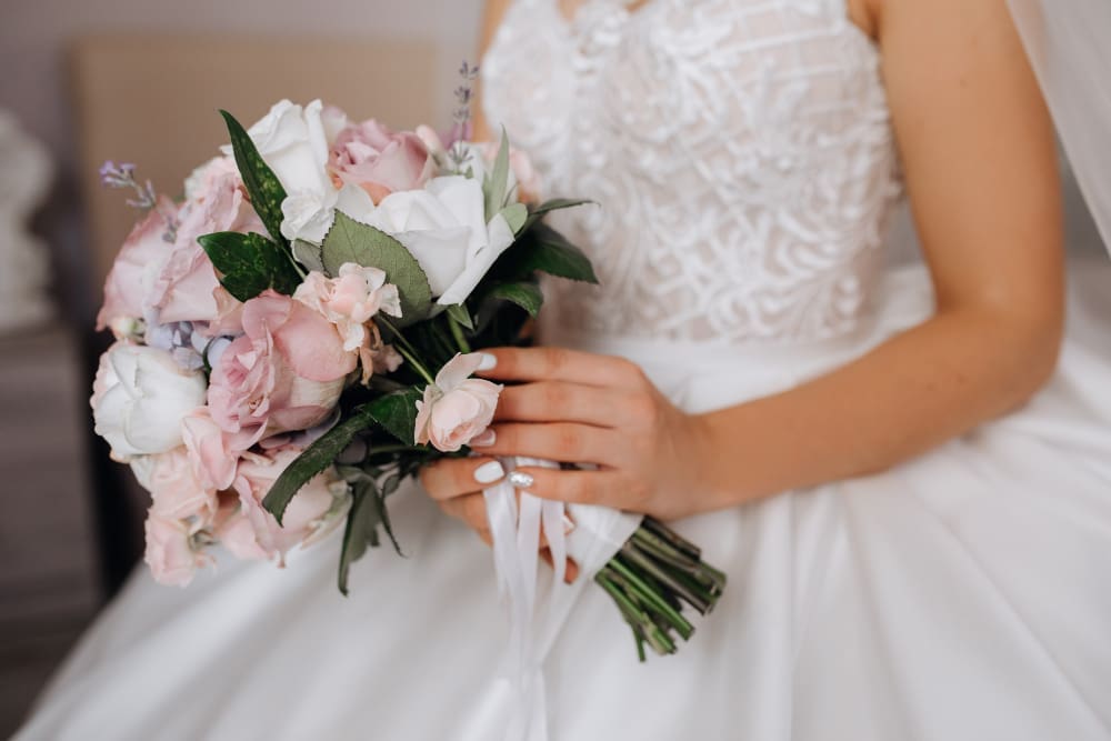 A bride in a white dress holds a bouquet of pink and white flowers with greenery and ribbon.