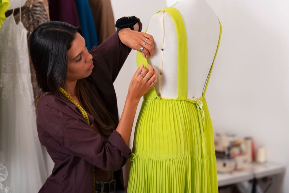 A person adjusts a bright yellow dress on a dress form in a workspace, wearing a measuring tape around their neck and using pins.
