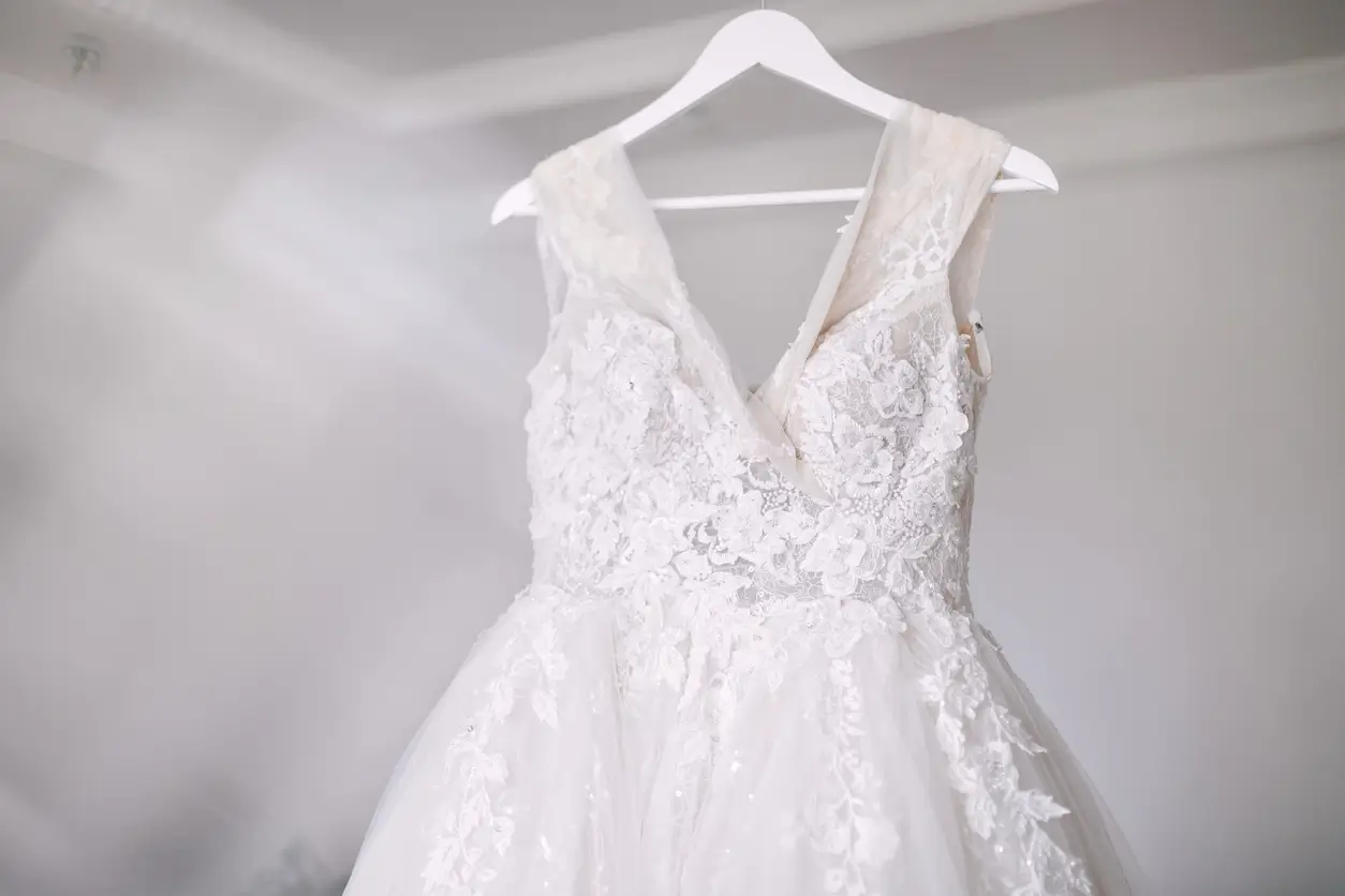 A white lace wedding dress with a tulle skirt hangs on a white hanger against a softly lit background.