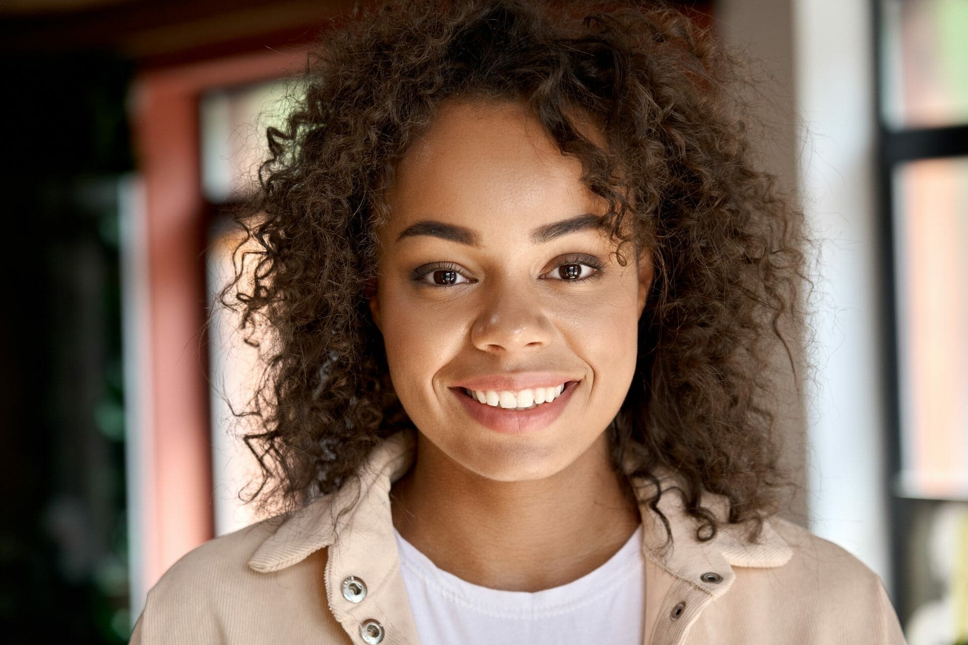 A young woman with curly hair smiling in front of a window.