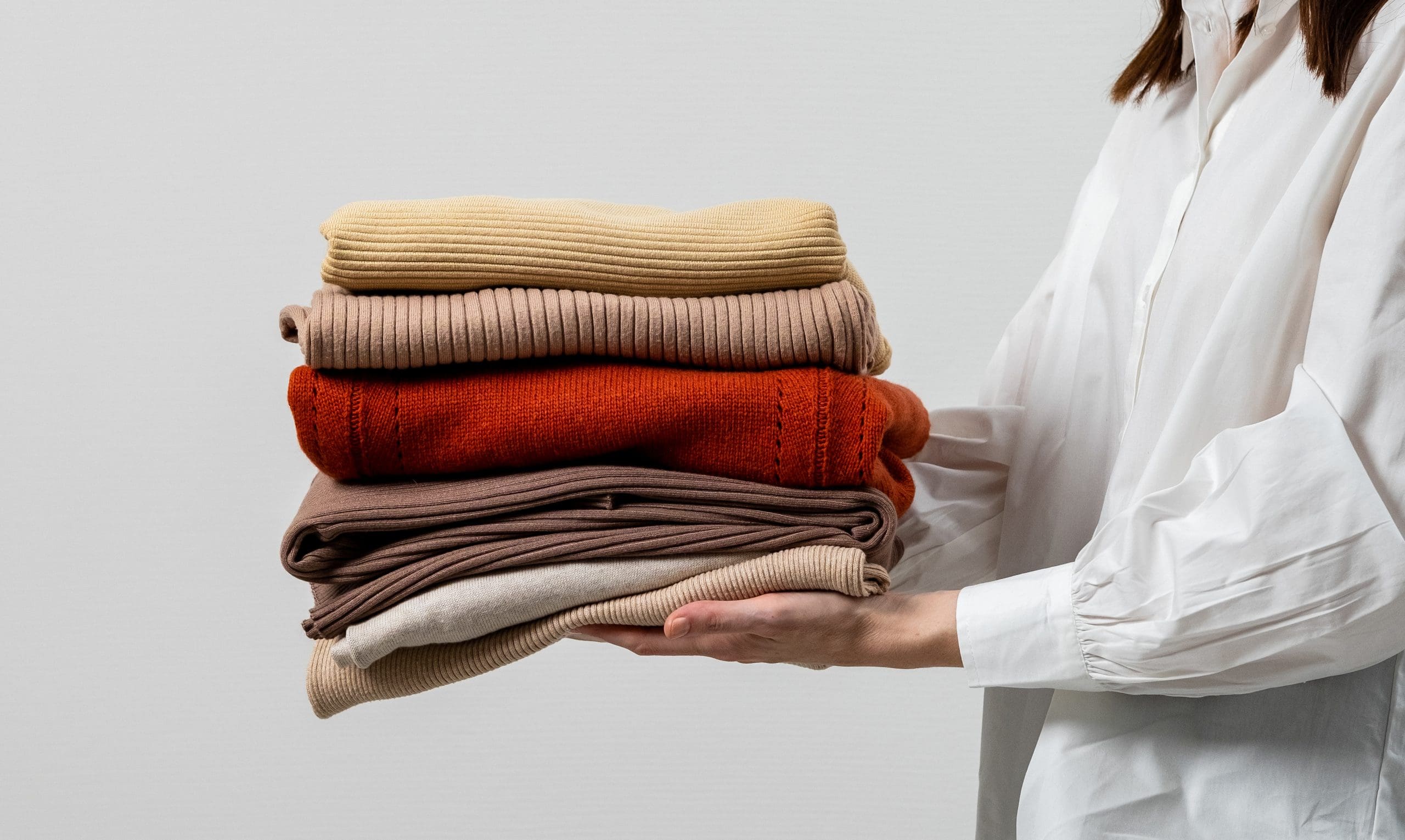 A person in a white shirt holds a stack of folded clothes in neutral and warm tones, including beige, orange, and brown.