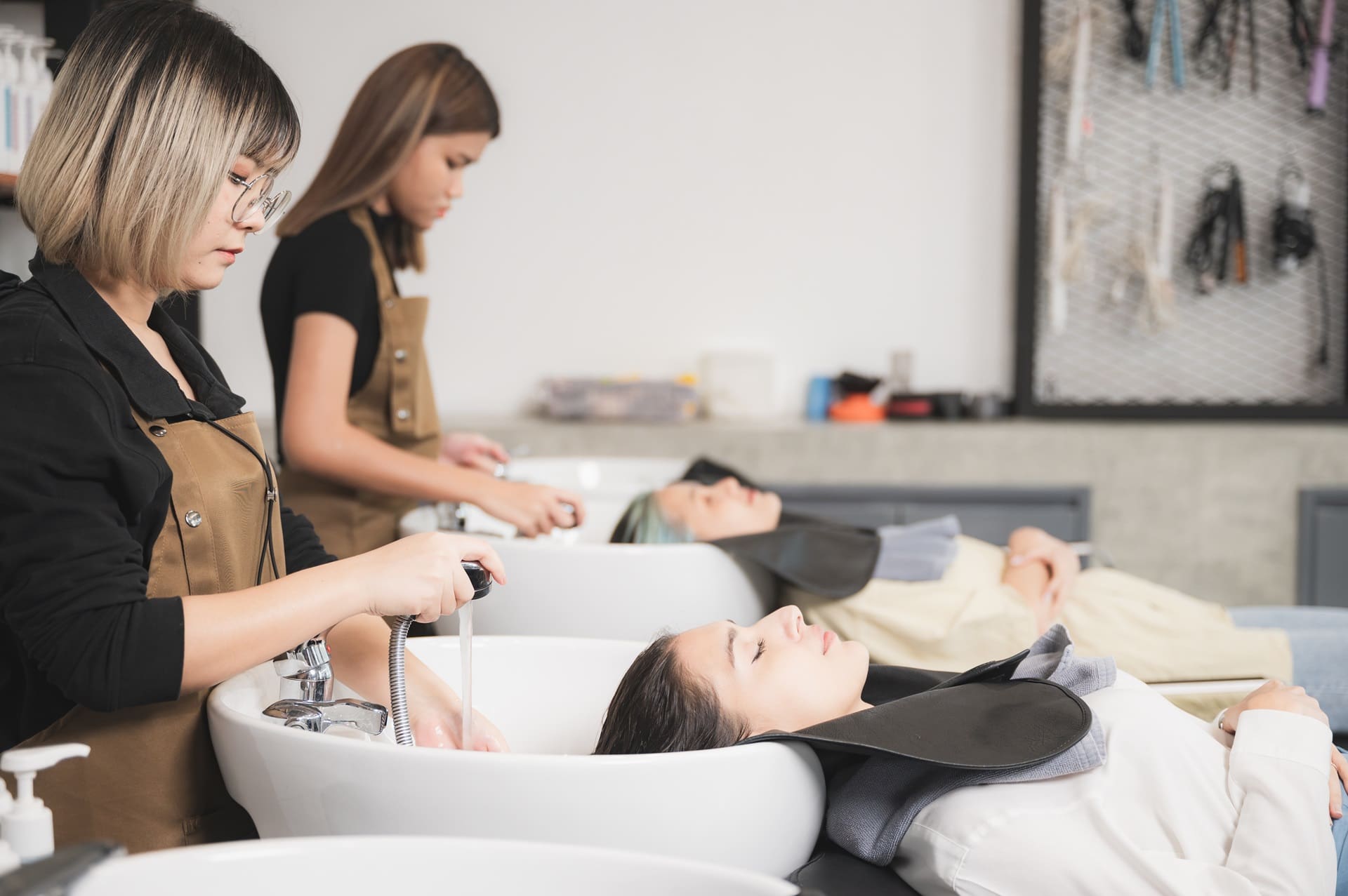 Two women lying with heads in sinks having their hair washed by stylists in a salon.