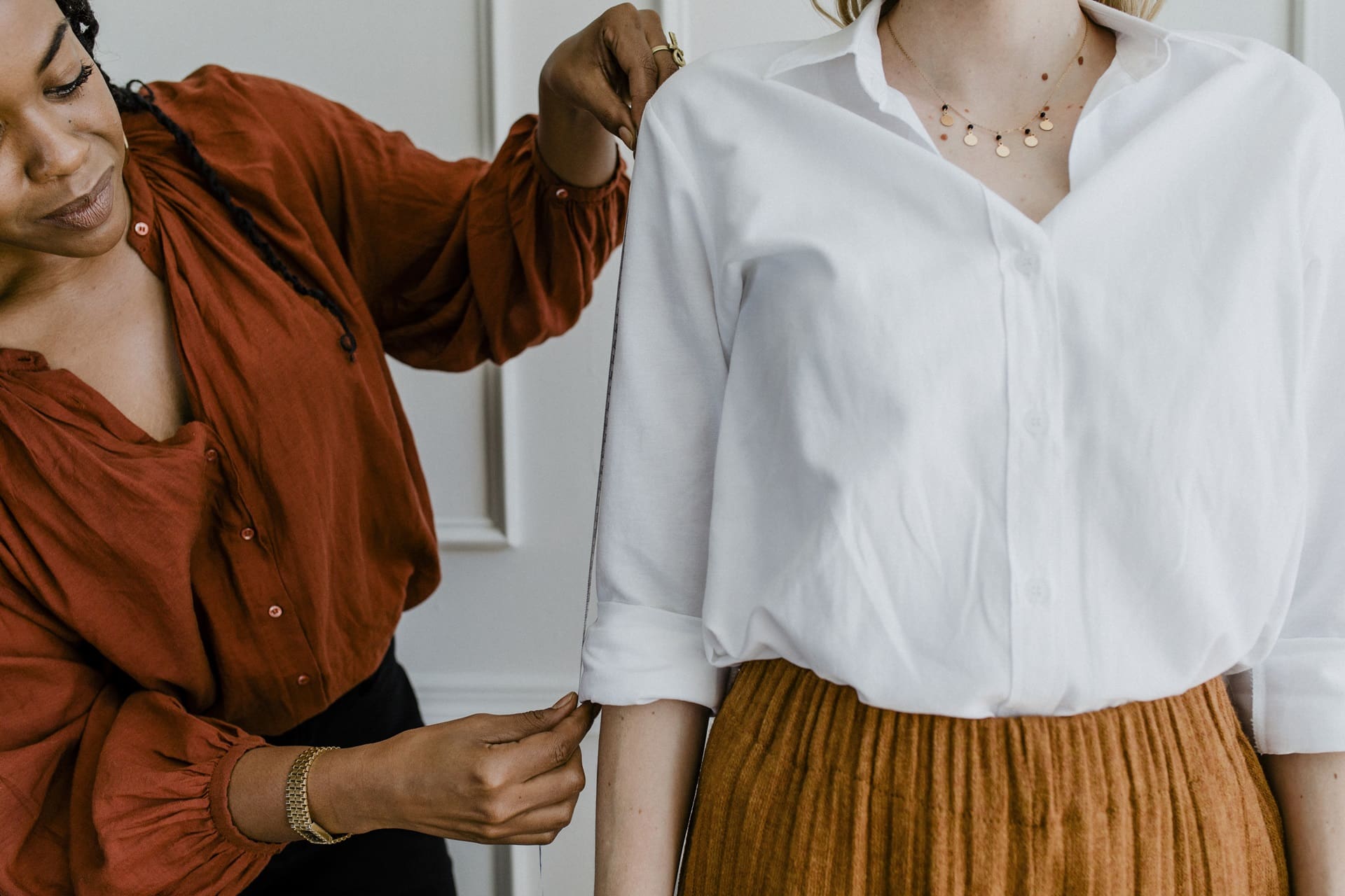 Person in a rust-colored shirt measures the sleeve length of a person wearing a white blouse and brown skirt.