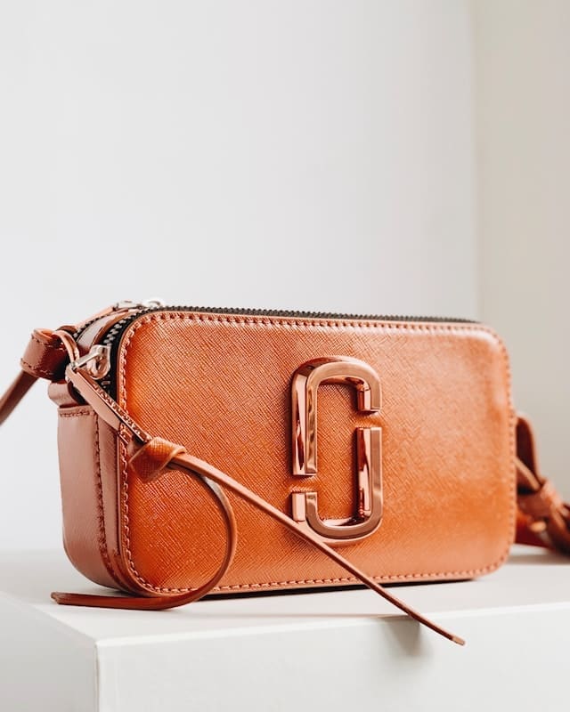 A small, rectangular, brown leather crossbody bag with a gold double-J logo and zipper, placed on a white surface against a light background.