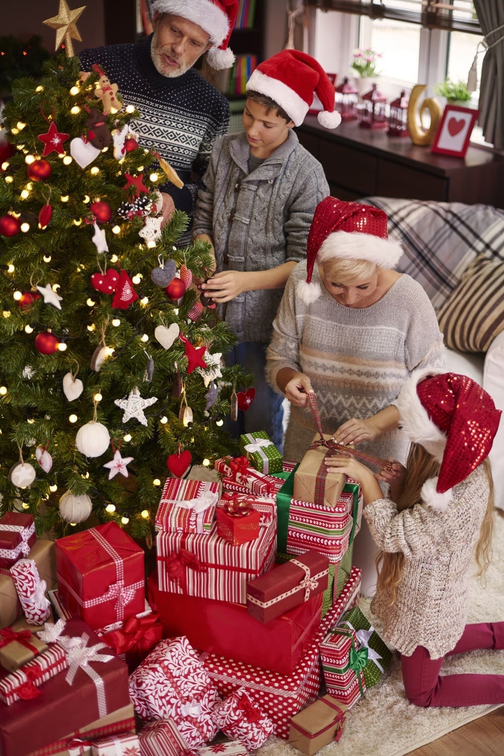 Four people in Santa hats decorate a Christmas tree and wrap gifts in a living room, surrounded by wrapped presents and festive decorations.
