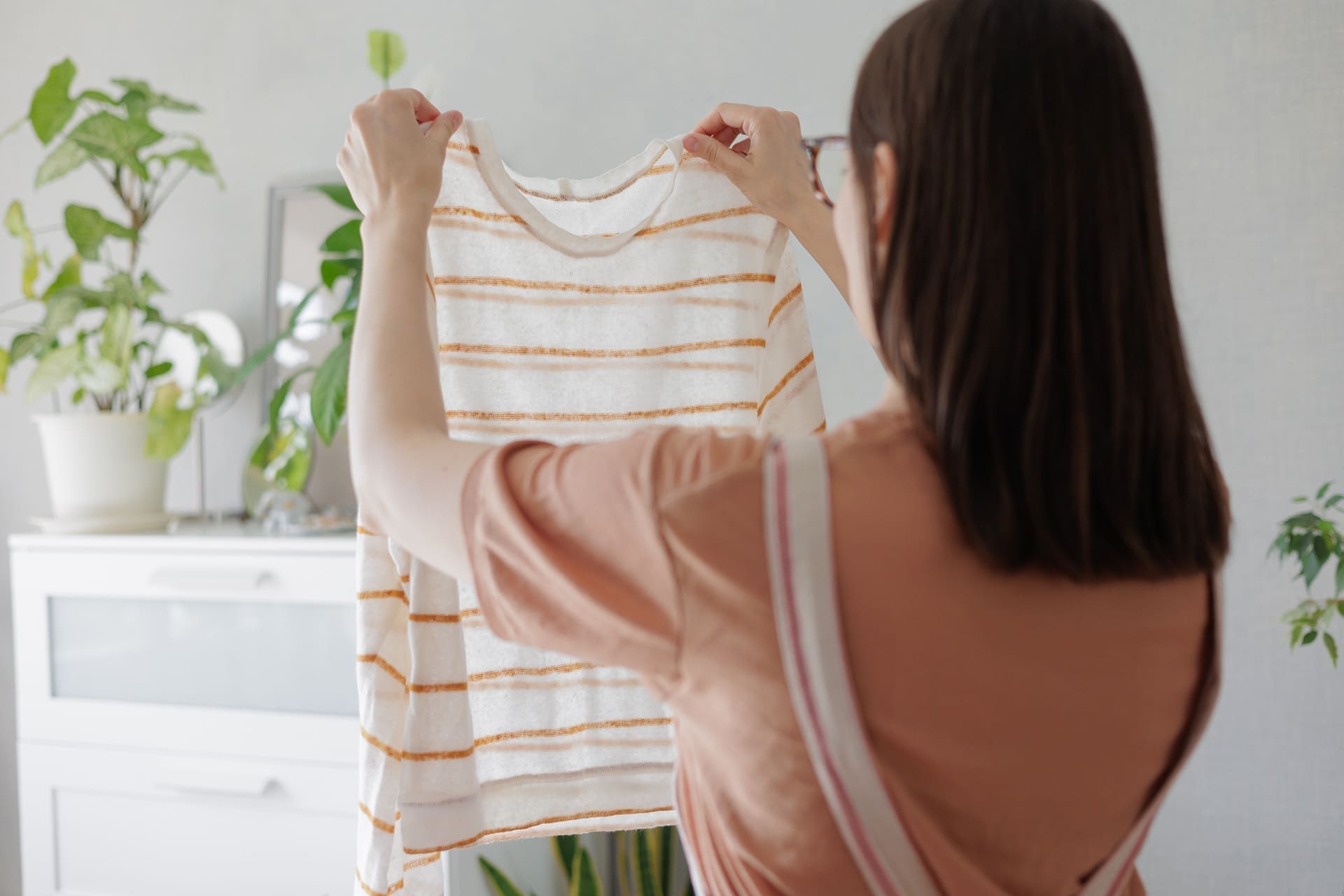 A person holds up a white and orange striped shirt, inspecting it, with indoor plants and a dresser visible in the background.