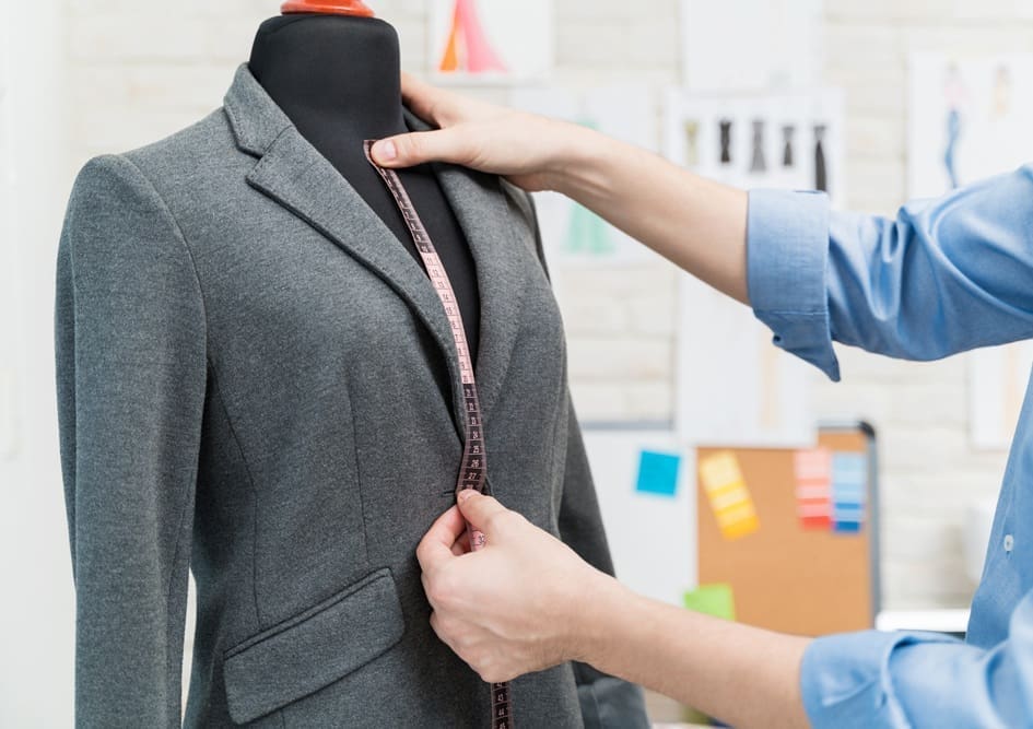 Person measuring a gray blazer on a black dress form with a measuring tape in a design studio.